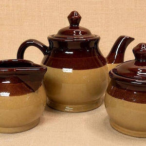 May include: A three-piece stoneware tea set in brown and tan. The set includes a teapot with a lid, a creamer, and a lidded sugar bowl. The teapot has a curved spout and a handle. The creamer has a handle and a spout. The sugar bowl has a lid with a knob.
