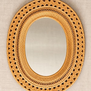 May include: An oval mirror featuring a light brown woven rattan frame. The frame consists of a central smooth band surrounded by an outer border of intricate, repeating braided patterns that create a textured, layered appearance.