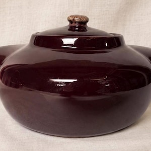 May include: A deep burgundy ceramic casserole dish with a matching lid and a small, textured knob. The dish has two side handles and a glossy finish. The background is a neutral fabric.