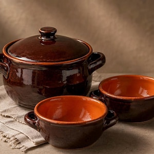 May include: A set of rustic brown ceramic cookware, including a lidded pot and two small bowls. The interior of the bowls is a vibrant orange. The cookware is placed on a beige linen cloth, suggesting a kitchen or dining setting.