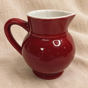 May include: A small, round, red ceramic pitcher with a handle and a spout. The interior is white, contrasting with the exterior's glossy finish. The pitcher sits on a small, circular base, showcasing a classic design.