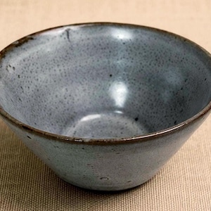 May include: A small, handmade ceramic bowl with a speckled, blue-gray glaze. The bowl has a slightly flared shape and a dark brown rim. The interior is smooth and glossy, while the exterior has a textured appearance.