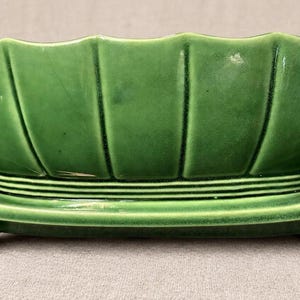 May include: A glossy, deep green ceramic planter featuring a scalloped, bowl-shaped upper section with vertical ridges. The base is rectangular with decorative scrolled ends and horizontal ribbed detailing connecting the bowl to the feet.