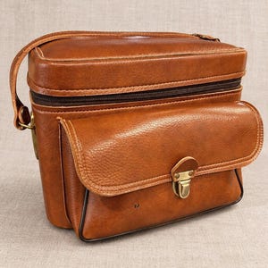 May include: A structured brown leather camera bag featuring a top zipper closure, a front flap pocket secured with a brass-toned metal clasp, and an adjustable shoulder strap. The bag has a textured finish and dark brown piping along the edges.