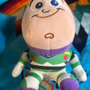 May include: A plush Buzz Lightyear doll, featuring a light purple head, blue eyes, and a smiling expression. The doll wears a green, white, and purple spacesuit. The toy is set against a colorful patterned background.