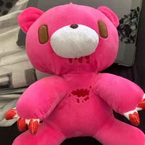 May include: A pink plush bear with brown ears and accents, a white muzzle, and red claws. The bear has a distressed appearance with red markings on its chest and mouth, giving it a unique, edgy aesthetic.