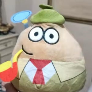 Może przedstawiać: Pluszowa zabawka w kształcie gruszki z dużymi oczami, zielonym kapeluszem i czerwoną krawatem. Ma fajkę i lupę. Zabawka jest beżowa, z czarno-białymi oczami i zielonym kapeluszem.