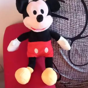 May include: A plush Mickey Mouse doll. The doll has a black body, black ears, and a white face. It wears red shorts with white buttons, white gloves, and yellow shoes. The doll is sitting on a red surface.