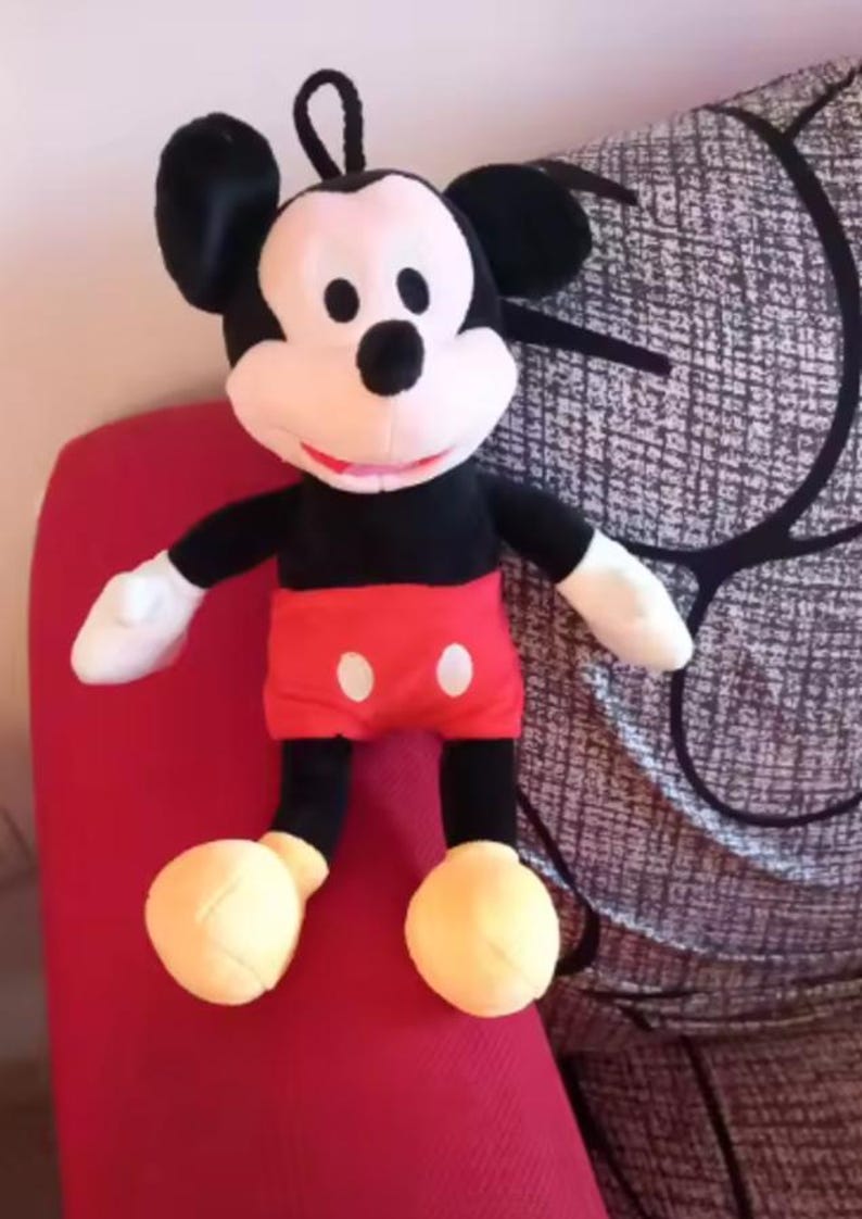 May include: A plush Mickey Mouse doll. The doll has a black body, black ears, and a white face. It wears red shorts with white buttons, white gloves, and yellow shoes. The doll is sitting on a red surface.