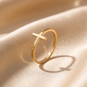 May include: A gold ring with a cross design. The ring is set against a soft, neutral-toned background, highlighting its simple yet elegant design. The cross is positioned on the top of the ring.