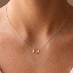 May include: A delicate gold-toned necklace featuring a thin, polished chain and a single, small circular pendant centered at the base. The metal has a warm, reflective finish, and the design consists of a simple, continuous loop suspended from the chain.