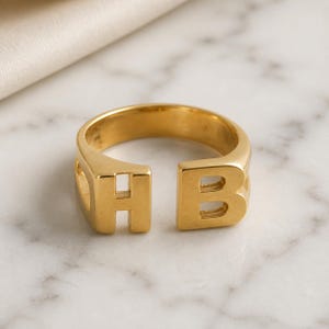 May include: A gold-toned open ring featuring two prominent block letters, H and B, positioned at the ends of the band. The ring has a smooth, polished finish and a wide, sturdy construction that creates a gap between the two letters.