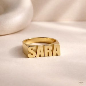 May include: A gold-colored ring with the name "SARA" in block letters. The ring is rectangular with a thick band. The letters are raised and stand out against the ring's surface. The ring is on a light-colored surface.