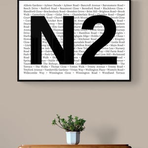 Könnte beinhalten: Gerahmter Druck mit einem großen schwarzen "N2" über einer Liste von Straßennamen. Eine kleine Topfpflanze in einem weißen Topf steht auf einer Holzkommode.