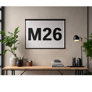 Op de afbeelding: Een zwart-wit print met de tekst "M26" in het midden. De achtergrond is gevuld met kleinere tekst. De print wordt getoond op een houten bureau met een lamp, boeken en decoratieve items.
