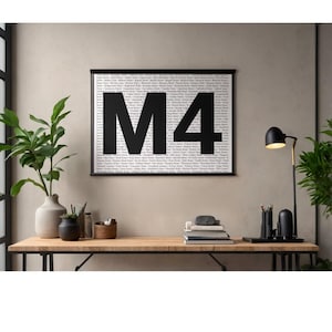 Op de afbeelding: Een ingelijste print met de grote zwarte letters "M4" in het midden. De achtergrond is gevuld met kleine tekst. De print wordt weergegeven op een houten bureau, samen met een lamp, boeken en diverse decoratieve items. De stijl is modern en minimalistisch.