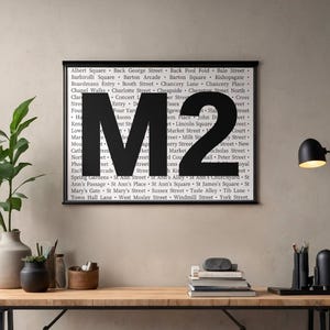 Op de afbeelding: Een ingelijste print met de zwarte letters "M2" op een achtergrond van tekst. De print staat op een houten bureau, met een lamp en planten. De print is ongeveer 60 cm breed.