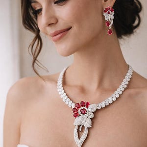 May include: A sparkling necklace and matching earrings set. The jewelry features a floral design with red gemstones and clear crystals. The necklace has a pendant with a teardrop-shaped red gemstone. The earrings have a similar floral design.