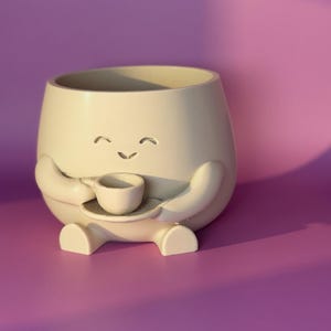 May include: A cream-coloured ceramic planter shaped like a smiling character, holding a small teacup and saucer. The planter has a rounded body, arms, and feet. The background is a solid purple.