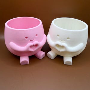 Puede incluir: Dos macetas sonrientes en forma de huevo, una rosa y otra blanca, cada una con brazos formando un corazón. Las macetas tienen pequeños pies y una abertura redonda en la parte superior para las plantas. El fondo es marrón liso.