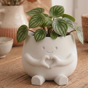 May include: A white ceramic mug with a black handle and the text "But First, Coffee" in a bold, black font. The mug is set against a blurred background, suggesting a cosy setting.