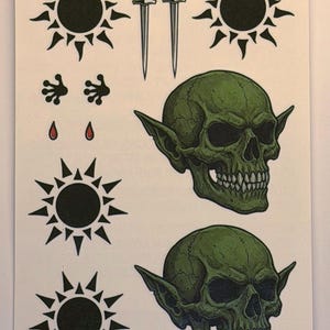 May include: A sheet of temporary tattoos featuring two green goblin skulls, four black sun symbols with varying ray shapes, two silver daggers, two small black frog-like footprints, and two small red teardrop shapes on a white background.