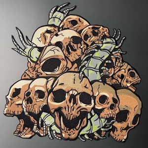 Op de afbeelding: Een geborduurde patch met een dichte stapel beige schedels met zwarte oogkassen en neusholtes, afgewisseld met verschillende gesegmenteerde, lichtgroene en grijze duizendpoten die in een gedetailleerd ontwerp over en tussen de schedels kruipen.