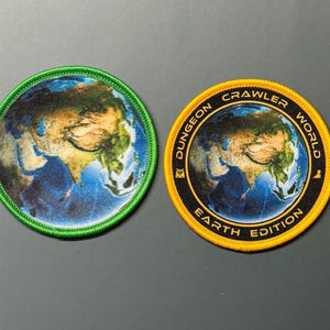 Pode incluir: Dois emblemas bordados circulares com uma vista de satélite da Terra. O emblema da esquerda tem uma borda verde brilhante. O emblema da direita tem uma borda preta com texto amarelo DUNGEON CRAWLER WORLD EARTH EDITION e uma borda amarela.