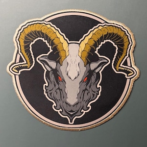 May include: A circular embroidered patch featuring a stylized goat head with large, curved golden-brown horns. The goat has a light gray face, dark gray fur, and piercing red eyes, set against a solid black circular background with a gold border.