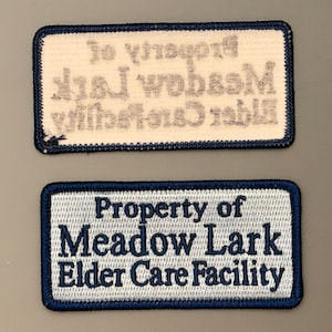 May include: Two rectangular embroidered patches with a white background and dark blue border. The text reads Property of Meadow Lark Elder Care Facility in dark blue thread. One patch shows the text clearly, while the other shows the reverse side.