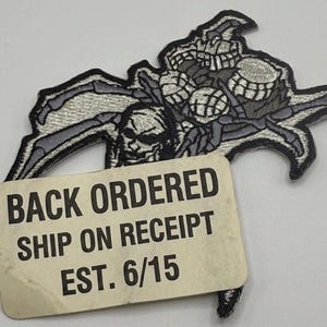May include: An embroidered patch featuring a grey and black skeletal figure with multiple limbs and a skull-like face. A rectangular paper tag is attached to the front with the black text: BACK ORDERED, SHIP ON RECEIPT, EST. 6/15.