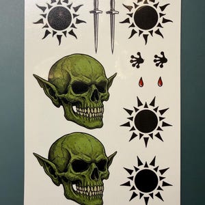 May include: A sheet of temporary tattoos featuring two green goblin skulls with pointed ears, two silver daggers, four black sun symbols with radiating points, two small black floral-like shapes, and two small red teardrop-shaped accents on a white background.