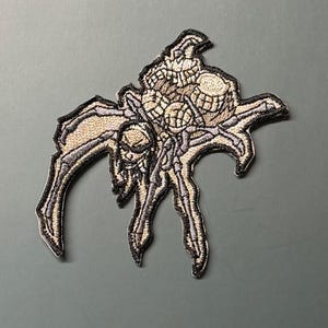 Könnte beinhalten: Ein gestickter Aufnäher, der eine stilisierte Spinne mit totenkopfartigem Kopf und segmentiertem Hinterleib zeigt. Das Design verwendet beigefarbene, hellbraune und dunkelgraue Fäden mit einem dicken schwarzen Rand für ein kontrastreiches Bild.