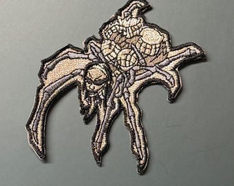 Könnte beinhalten: Ein gestickter Aufnäher, der eine stilisierte Spinne mit totenkopfartigem Kopf und segmentiertem Hinterleib zeigt. Das Design verwendet beigefarbene, hellbraune und dunkelgraue Fäden mit einem dicken schwarzen Rand für ein kontrastreiches Bild.