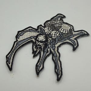 May include: Embroidered patch featuring a stylized spider design with a skull-like head and multiple smaller skulls. The patch is primarily gray with black and white accents, creating a detailed and gothic aesthetic. The patch is likely for clothing or accessories.