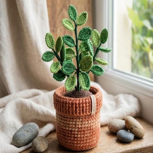 Może przedstawiać: Ręcznie robiona roślina na szydełku w doniczce w kolorze terakoty. Roślina ma zielone liście i brązową podstawę, przypominającą ziemię. Doniczka ma teksturowany wzór i małą etykietę z napisem "crochet & co.".