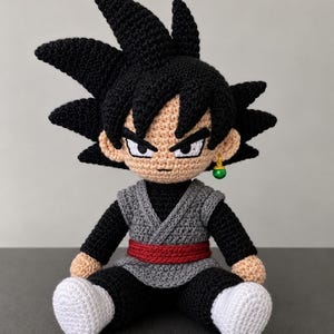 Puede incluir: Muñeco de ganchillo de un personaje con pelo negro puntiagudo, piel morena y expresión seria. El muñeco lleva un chaleco gris, pantalones negros, un cinturón rojo y zapatos blancos. Un pendiente verde cuelga de una oreja.