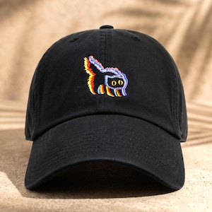 May include: A black baseball cap featuring an embroidered graphic of a cat with a glitch effect. The cat design displays vibrant, layered colors including red, blue, and yellow, with two large, round, yellow eyes centered on the front panel.