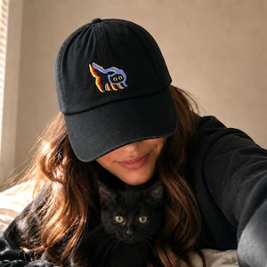 May include: A black baseball cap featuring an embroidered graphic of a small cat with a colourful, multi-toned outline. The cap has a curved brim and a structured crown, showcasing a detailed stitched design of a feline figure in vibrant hues.