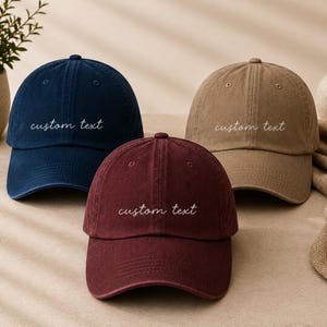 Könnte beinhalten: Drei Baseballkappen in Marineblau, Kastanienbraun und Hellbraun, die jeweils mit dem Schriftzug custom text in weißer Schreibschrift auf der Vorderseite bestickt sind. Die Kappen haben eine strukturierte Krone, einen gebogenen Schirm und einen Knopf.