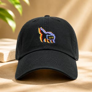 May include: Black baseball cap featuring a colourful embroidered cat design. The cat has a glitch effect with red, yellow, and blue outlines. The cap has a curved peak and a fabric strap closure.