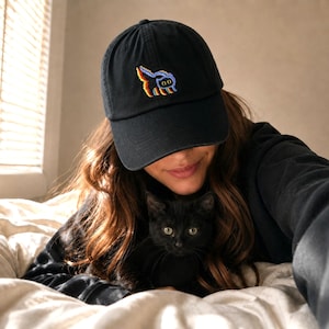 May include: Black baseball cap with a colorful embroidered design. The cap is worn by a person holding a black cat. The cat has green eyes and is looking at the camera. The background is a bedroom with a window.