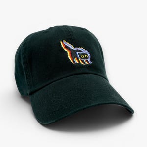 May include: A dark green baseball cap with a curved brim. The cap features a colorful embroidered design of a cartoon skunk on the front. The skunk has a rainbow-colored tail and a pair of eyes. The cap is made of a soft, durable fabric.
