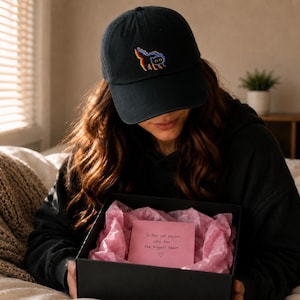 May include: A black baseball cap with a colorful embroidered cat design. The person is holding a black gift box with pink tissue paper and a pink card that reads, "To the cat person who has the biggest heart".