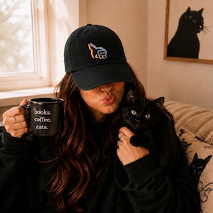 May include: A black baseball cap with a colorful graphic and a black mug with the words "books. coffee. cats." A black cat is held in the arms of a person. A framed cat silhouette art piece hangs on the wall.