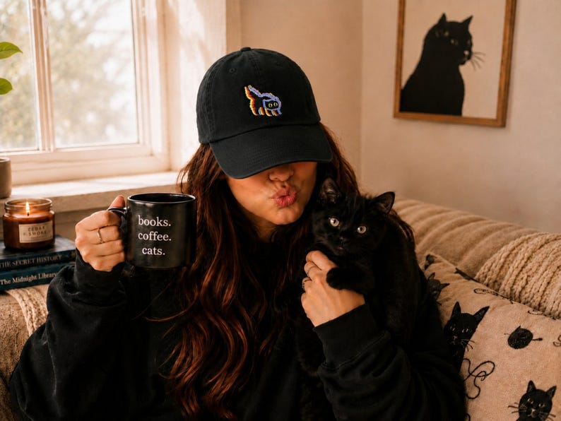 May include: A black baseball cap with a colorful graphic and a black mug with the words "books. coffee. cats." A black cat is held in the arms of a person. A framed cat silhouette art piece hangs on the wall.