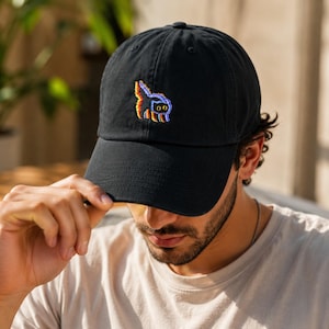 May include: Black baseball cap with a curved brim. The cap features a colorful embroidered design on the front. The design appears to be a stylized cat or creature. The cap is made of a soft material.