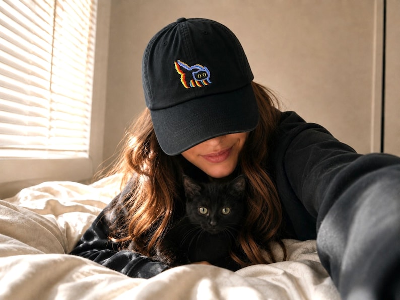 May include: A black baseball cap featuring an embroidered graphic of a small cat with a colourful, multi-toned outline. The cap has a curved brim and a structured crown, showcasing a detailed stitched design of a feline figure in vibrant hues.