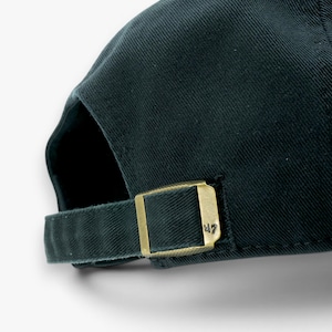 May include: Dark green baseball cap with a curved brim. The adjustable strap features a gold-colored metal buckle with the number "47" engraved on it. The cap is made of a textured fabric.