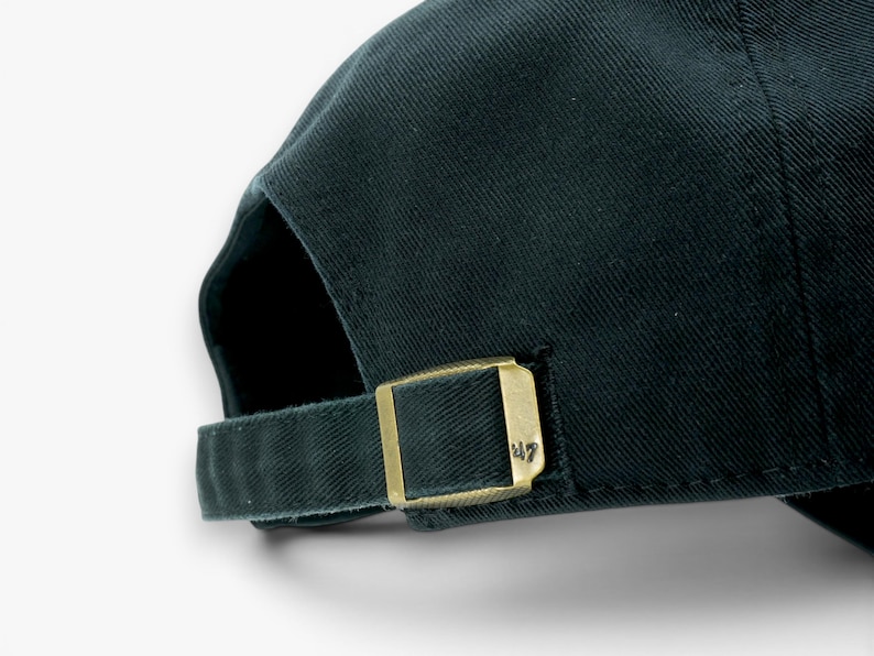 May include: Dark green baseball cap with a curved brim. The adjustable strap features a gold-colored metal buckle with the number "47" engraved on it. The cap is made of a textured fabric.